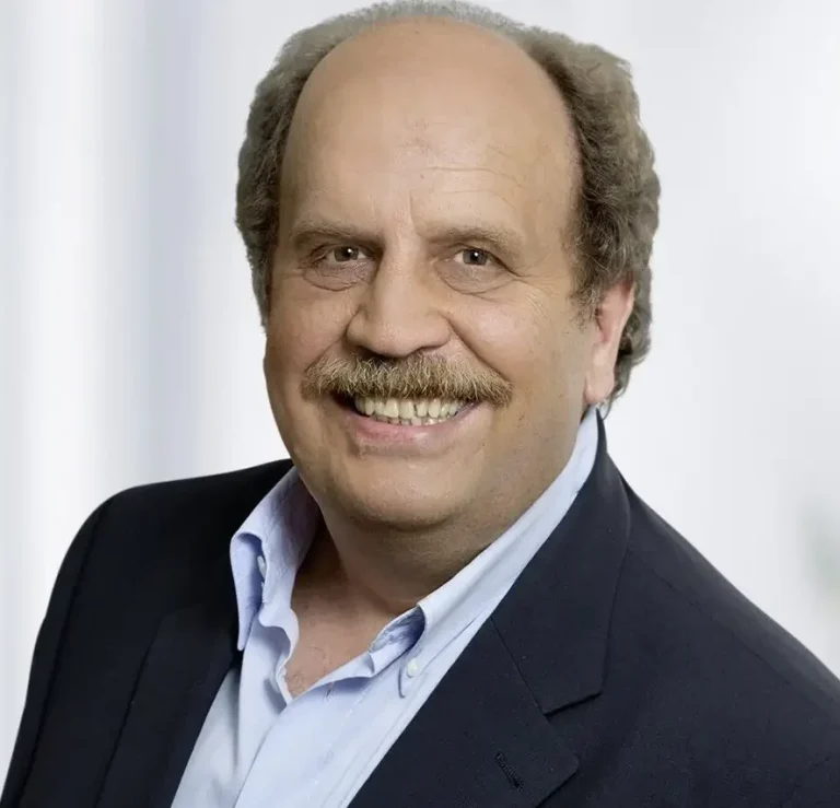 Warum CaPe Consult Lächelnder Mann mit dunkler Blazer und hellblauer Hemd, vor neutralem Hintergrund.