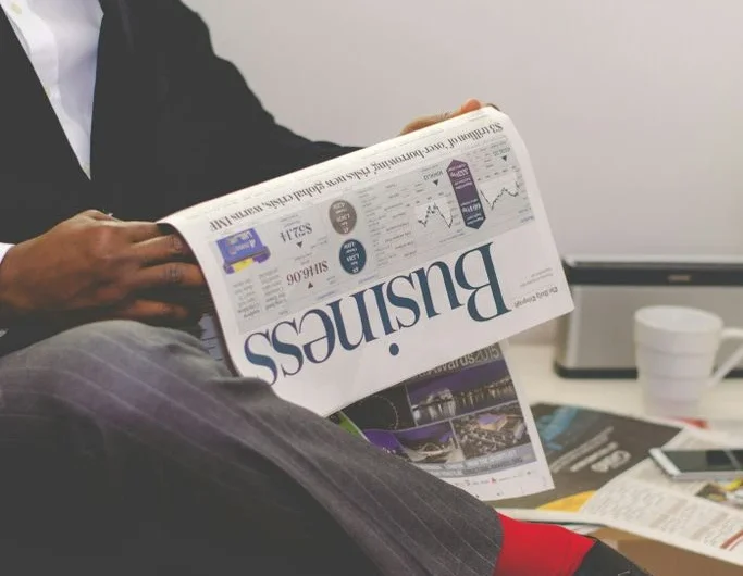 Betriebswirtschaftliche Beratung Mann in Anzug liest eine Zeitung, neben ihm stehen Pflanzen und ein Kaffeebecher.