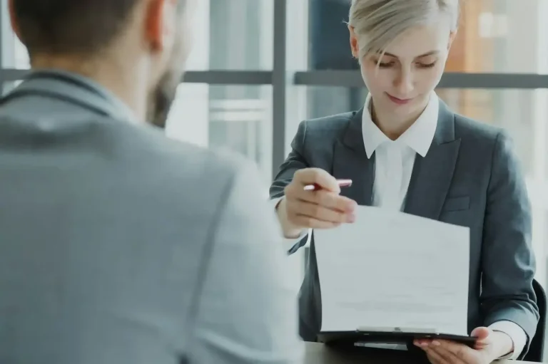 AVGS-Schein Frau in Anzug reicht Dokument an einen Mann in grauem Anzug.